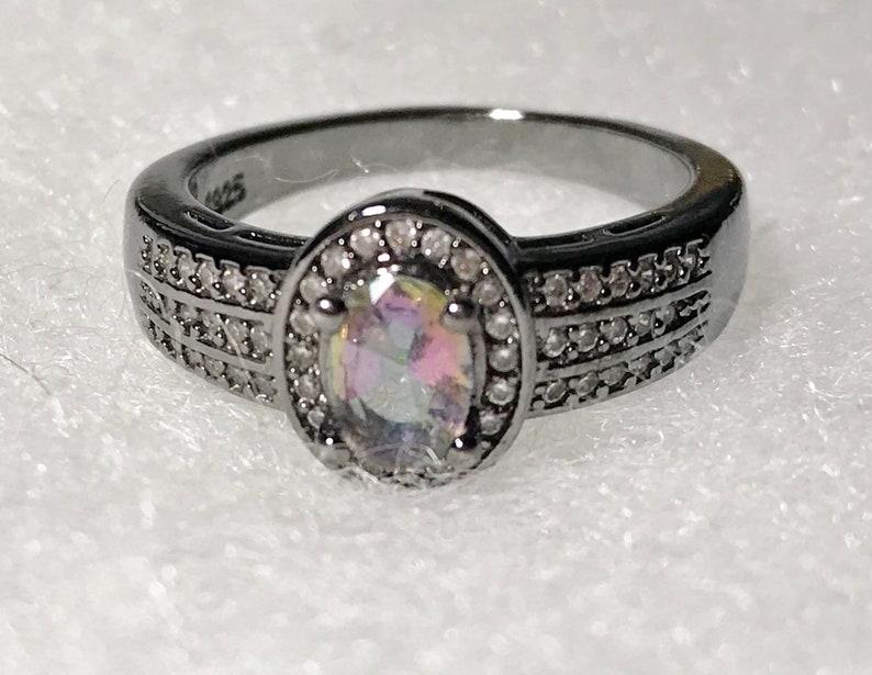 May include: A black ring with a large oval-shaped iridescent gemstone surrounded by a halo of small clear stones. The band of the ring is decorated with two rows of small clear stones.