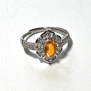 May include: A silver ring with a large oval orange gemstone surrounded by smaller clear gemstones. The ring has a delicate floral design.