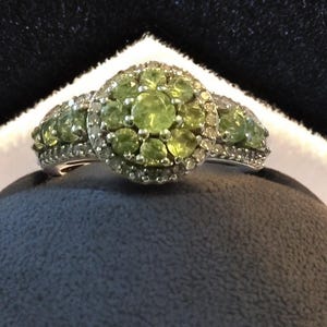 May include: A silver ring with a cluster of round, light green gemstones in the center, surrounded by smaller clear stones. The ring has a halo design with additional green stones on each side.