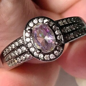 May include: A black ring with a large oval-shaped iridescent gemstone surrounded by a halo of smaller clear gemstones. The band has two rows of smaller clear gemstones.