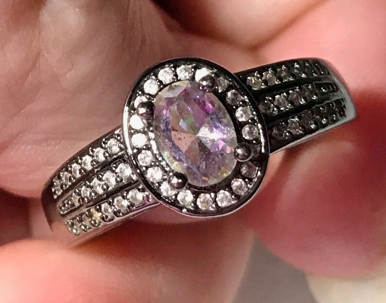 May include: A black ring with a large oval-shaped iridescent gemstone surrounded by a halo of smaller clear gemstones. The band has two rows of smaller clear gemstones.