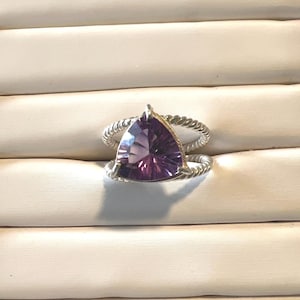 May include: A silver ring featuring a triangular, faceted purple gemstone. The ring's band has a twisted rope design. The gemstone is a deep purple color and is the focal point of the ring. The ring is displayed on a white surface.