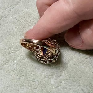 May include: A gold ring with a large purple gemstone surrounded by smaller clear gemstones. The ring is set in a delicate, intricate design.