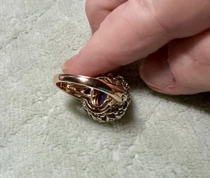 May include: A gold ring with a large purple gemstone surrounded by smaller clear gemstones. The ring is set in a delicate, intricate design.