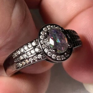 May include: Black ring with a large oval-shaped iridescent gemstone surrounded by a halo of smaller clear gemstones. The band is decorated with a row of smaller clear gemstones.