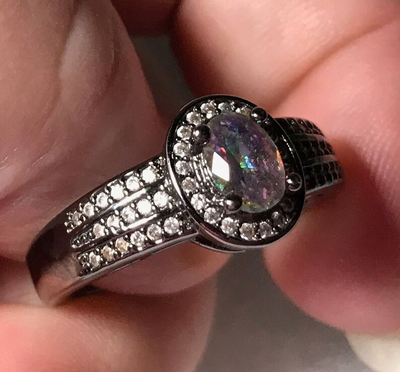 May include: Black ring with a large oval-shaped iridescent gemstone surrounded by a halo of smaller clear gemstones. The band is decorated with a row of smaller clear gemstones.