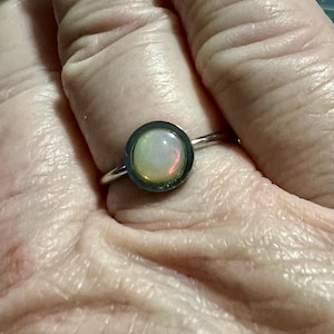 May include: A silver ring with a round opal gemstone set in a black bezel. The opal has a white, yellow, and pink iridescent sheen.