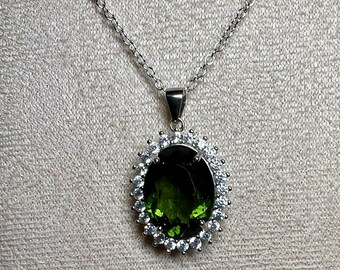 May include: A silver chain necklace with a large oval pendant featuring a dark green gemstone surrounded by a halo of smaller clear gemstones.