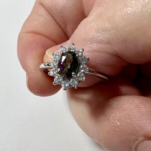 Peut inclure: Une bague en argent avec une grande pierre précieuse ovale irisée entourée de petites pierres précieuses claires.