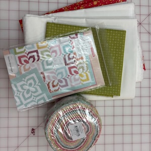 Könnte beinhalten: Ein Quilt-Zubehör-Set bestehend aus einem Kaleidoscope-Musterheft in einer Klarsichthülle, einer in Plastik verpackten Rolle mit bunten Stoffstreifen, einem gefalteten grünen Stoff mit weißem Gittermuster sowie gestapelten weißen und roten Blumenstoffen.