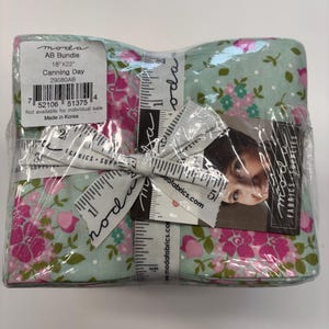May include: A fabric bundle wrapped in clear plastic, with a floral pattern of pink flowers and green leaves on a light blue background. The bundle is tied with a ribbon printed with measurements in inches and the text "moda fabrics". The label reads "Canning Day". The bundle measures 45.7cm x 55.9cm.