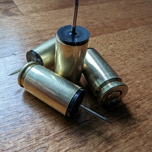 May include: Three brass bullet casings with black tops and metal pins protruding from the tops. The casings are arranged in a triangular shape on a wooden surface.