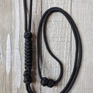 May include: Black rope halter with a braided handle and a loop at the top. The halter features two decorative knots near the end of the rope. The background is a weathered wood surface.