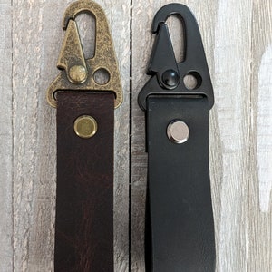 May include: Two leather keychains with metal clips. One keychain is brown with a brass clip and the other is black with a black clip.