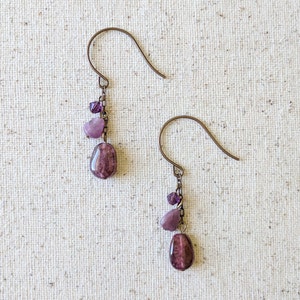 May include: A pair of dangle earrings with purple gemstone beads and small purple glass beads hanging from a bronze chain.
