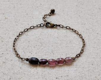May include: A delicate bracelet with a chain made of antique gold metal and a row of oval-shaped gemstones in shades of pink and purple.