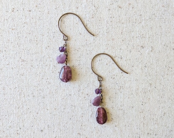 May include: A pair of dangle earrings with a dark bronze hook and chain. Each earring features a large, oval, dark purple bead, two smaller light purple beads, and several tiny purple beads. The earrings are displayed on a textured, off-white surface.