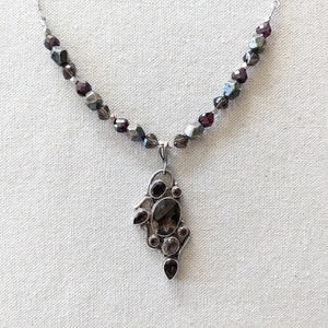 May include: A silver necklace with a pendant featuring a cluster of dark brown and purple gemstones. The necklace is made with small, faceted beads in shades of gray and purple.