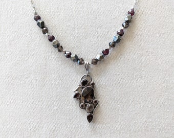 May include: A silver necklace with a pendant featuring a cluster of dark brown and purple gemstones. The necklace is made with small, faceted beads in shades of gray and purple.