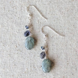 May include: A pair of dangle earrings with silver-colored hooks. Each earring features a chain with small blue beads, dark purple oval beads, and a large, oval, pale green stone. The earrings are displayed on a textured, neutral-colored surface.