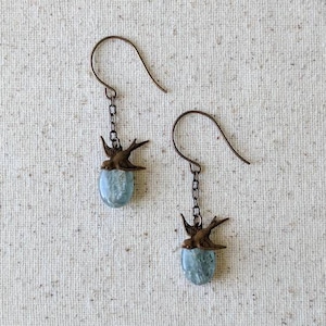 May include: A pair of earrings featuring a bird design. Each earring has a bronze-colored bird charm perched above a light blue, oval-shaped stone. The earrings hang from bronze-colored ear wires.