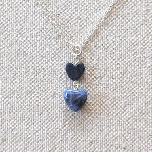May include: A silver chain necklace with two heart-shaped pendants. The top heart is dark blue, and the bottom heart is a lighter blue with a marbled appearance. The necklace is displayed against a neutral background.