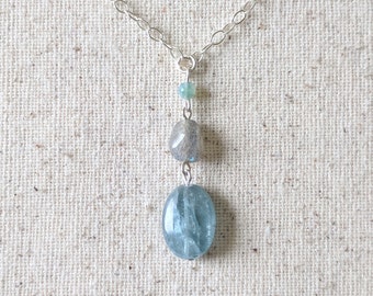 May include: A silver chain necklace with a pendant featuring three gemstones. The pendant has a small, round, light green bead at the top, followed by a gray, faceted stone, and a large, oval, light blue stone at the bottom. The necklace is displayed on a beige fabric.