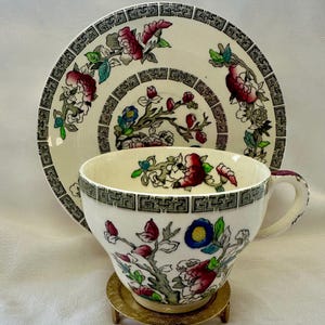 Puede incluir: Un juego de taza y platillo de té vintage. La taza y el platillo de porcelana blanca presentan un diseño floral con detalles en rojo, azul y verde. El borde de la taza y el platillo tiene un borde geométrico negro y gris. La taza está sobre un pequeño soporte dorado.