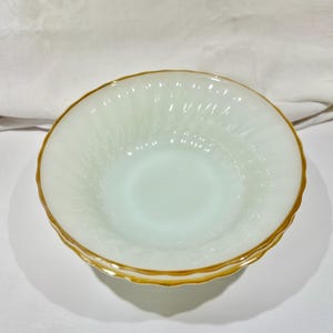 Può includere: Due ciotole di vetro bianco con bordo dorato. Le ciotole hanno un motivo strutturato all'interno e un bordo smerlato. Le ciotole sono su una superficie bianca.