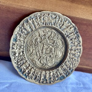 May include: A round, antique-style brass plate with an intricate relief design. The plate features a central scene with figures and a detailed border with additional figures and floral elements. The plate has a slightly tarnished appearance.
