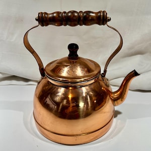 May include: A copper tea kettle with a wooden handle and a black knob on the lid. The kettle has a rounded body and a curved spout. The handle is made of dark brown wood. The kettle is made of shiny copper.