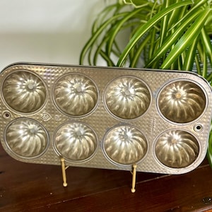 Könnte beinhalten: Vintage silberfarbene Bundt-Kuchenform mit acht einzelnen Formen. Die Form hat eine strukturierte Oberfläche und ein dekoratives Design in jeder Form. Die Form wird von einem goldfarbenen Ständer getragen, vor einem Hintergrund aus grünem Laub.