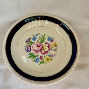 Puede incluir: Un plato redondo de color crema con un borde azul marino y un diseño floral. El centro del plato presenta un ramo de flores rosas, moradas, azules y amarillas. El borde tiene una textura de cuentas.