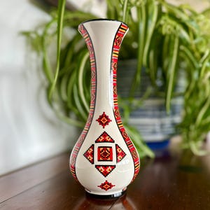 Puede incluir: Un jarrón de cerámica de color crema con un cuello delgado y una base bulbosa. La superficie está decorada con intrincados patrones geométricos en rojo, negro y amarillo, con motivos de diamantes y cruces que envuelven todo el recipiente.