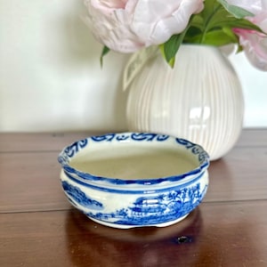 May include: A small, round ceramic bowl featuring a white base with intricate blue hand-painted landscape scenes depicting pagodas, trees, and mountains. The rim has a scalloped edge with a decorative blue pattern, and the interior is plain white.