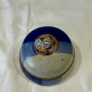 Op de afbeelding: Een keramische kom met deksel, in blauwe en bruine tinten. Het deksel is versierd met een bloemmotief. De ronde kom heeft een gespikkelde textuur, ideaal om te serveren of op te bergen.