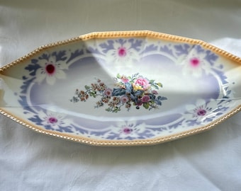 May include: Oval porcelain serving dish with a floral design. The dish has a light purple and white background with a gold trim. The centre of the dish has a bouquet of pink, yellow, and purple flowers.