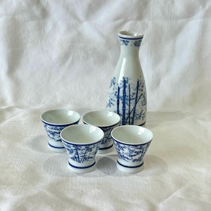 May include: A white ceramic sake set with a blue bamboo design. The set includes a tall bottle and four small cups. The bamboo pattern is repeated on the bottle and cups, creating a cohesive look. The set is ideal for serving sake.