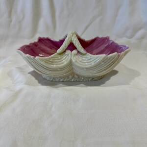 May include: A decorative, shell-shaped dish with a handle. The dish has a cream-coloured exterior and a vibrant pink interior. The handle is cream-coloured and the dish has a scalloped edge.