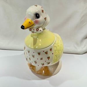 Puede incluir: Un tarro de galletas de cerámica con forma de pato. El pato tiene un pico amarillo, mejillas rosadas y una cabeza blanca con detalles florales rojos. El cuerpo es amarillo con un delantal estampado floral y una espalda texturizada. La base es marrón.