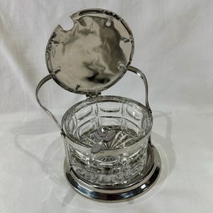Könnte beinhalten: Ein Vintage-Zuckerdose mit einem transparenten, geschliffenen Glasbehälter und einem silberfarbenen Metallrahmen, Deckel und Griff. Der Deckel ist geöffnet und gibt den Blick ins Innere frei. Die Schale steht auf einem runden Sockel.