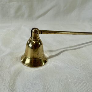 May include: A vintage brass candle snuffer with a bell-shaped head and a long handle. The candle accessory is made of shiny, aged brass and sits on a white, textured surface. The handle extends horizontally from the bell.