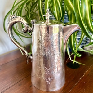 May include: A silver-coloured metal coffee pot featuring an ornate, engraved scrollwork pattern on the front, a curved handle with decorative detailing, a hinged lid with a small finial, and a tapered spout extending from the side of the body.