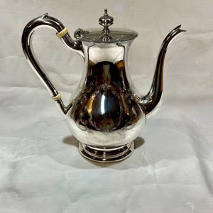 Puede incluir: Una cafetera plateada con un asa curva y un pico largo y delgado. La cafetera tiene detalles ornamentados alrededor del cuerpo y un pequeño adorno decorativo en la tapa. La cafetera se asienta sobre una base redonda.