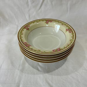 May include: A stack of five white ceramic bowls. Each bowl has a cream-coloured rim with a gold border and a floral pattern in pink and green. The bowls are stacked on top of each other.