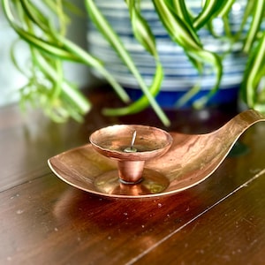 Puede incluir: Un candelero de cobre con forma de hoja. Tiene una pequeña copa circular para la vela y un mango largo y curvado. El cobre tiene un tono marrón rojizo cálido y descansa sobre una superficie de madera.