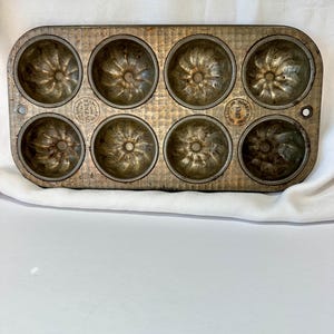 Könnte beinhalten: Vintage Bundt-Kuchenform aus Metall mit acht einzelnen Formen. Die Form hat eine rechteckige Form mit abgerundeten Kanten und einem abgenutzten, gealterten Aussehen. Jede Form weist ein detailliertes, geriffeltes Design auf. Die Form trägt die Aufschrift "Ovenex" und "1387".