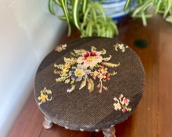May include: A small, round footstool featuring a dark brown needlepoint fabric top with a central floral motif in shades of pink, yellow, and green. The stool is supported by three short, turned wooden legs with a worn, light brown finish.