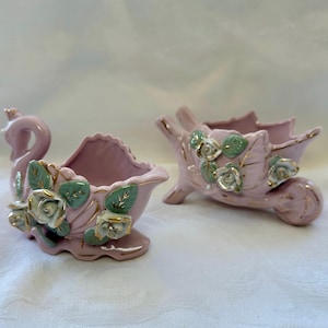 May include: Two pink ceramic decorative planters. One is swan-shaped, the other is a wheelbarrow. Both are decorated with white and gold roses and green leaves. The planters have a glossy finish and are set against a neutral background.