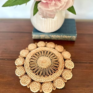 May include: A circular woven placemat with a floral pattern. The placemat is made from natural fibres, with a dark brown central design and outer ring. It sits on a dark wooden surface, with a white vase and pink flowers in the background.